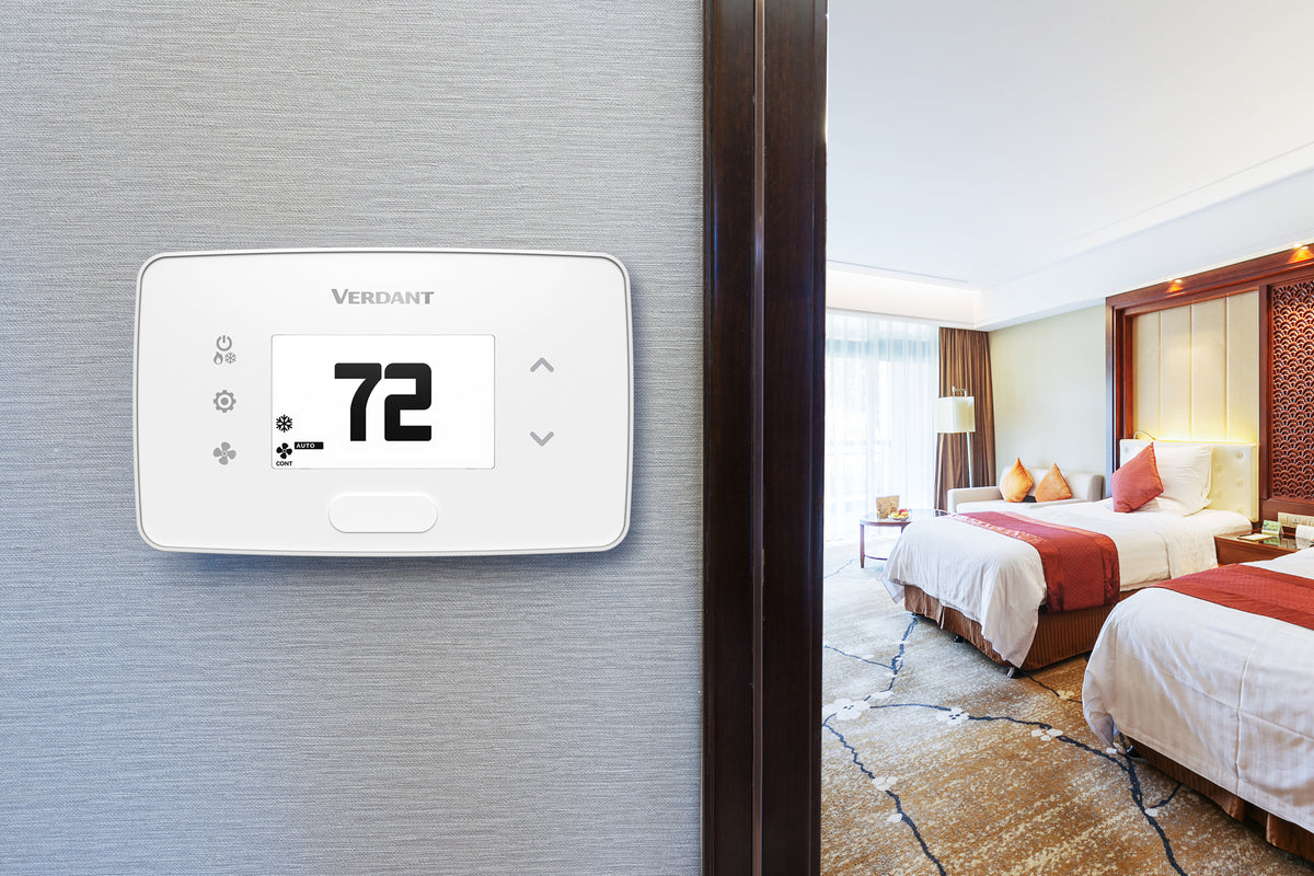 A white digital thermostatic control panel by Verdant mounted on a textured gray wall, with a hotel room visible in the background through an open doorway.